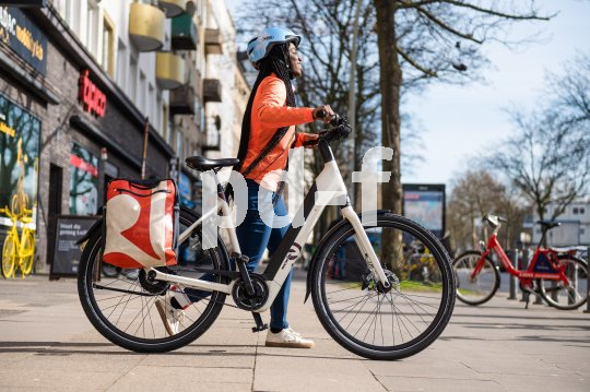 Eine Frau schiebt ein E-Bike über einen Gehweg vor einer Häuserzeile mit Geschäften.