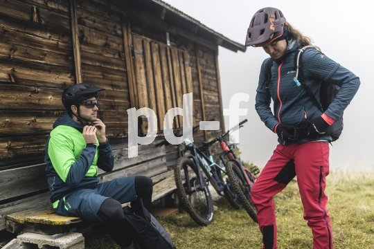 Ein Mann und eine Frau machen sich vor einer Holzhütte startklar zum Radfahren. An der Hütte lehnen zwei Mountainbikes.
