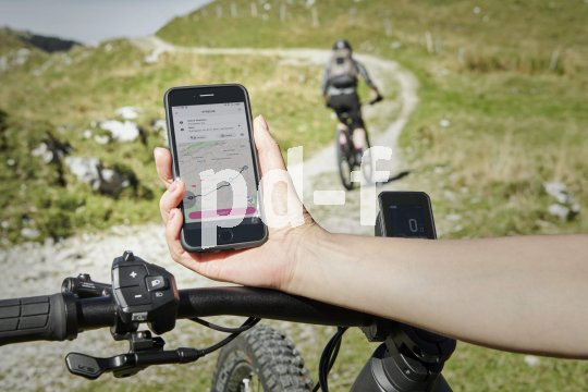 Eine Person hät ein Smartphone mit Karten-App darauf über den Lenker eines E-Bikes. Im Hintergrund fährt eine Person einen Wirtschaftsweg entlang.