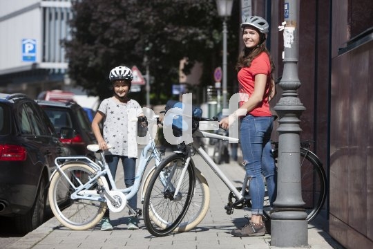 Ein Mädchen und eine Frau stehen mit Fahrrädern auf einem Bürgersteig. Beide tragen Helm.