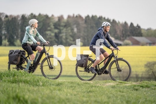 Eine Frau und ein Mann fahren auf E-Bikes zwischen Feldern und Wiesen entlang.