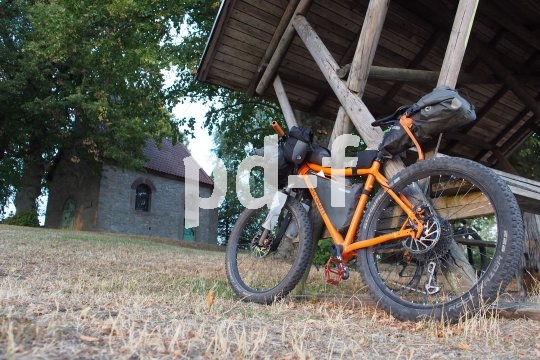 Ein mit Gepäck beladenes, orangefarbenes Mountainbike lehnt an einem hölzernen Unterstand auf einer Wiese vor einer Kapelle.