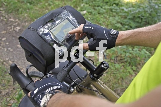 Eine Person tippt mit dem Finger auf das Display eines Smartphones oder Navigationsgeräts in einer Klarsichthülle auf dem Deckel einer Lenkertasche eines Fahrrades.