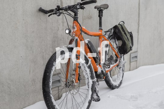 Ein orangefarbenes E-Bike mit Schnee in den Rädern und am Rahmen steht an eine Wand gelehnt.
