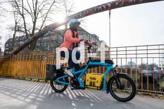 Eine Frau fährt auf einem blauen Lastenrad mit einem gelben Getränkekasten auf der Ladefläche über eine Brücke in der Stadt.