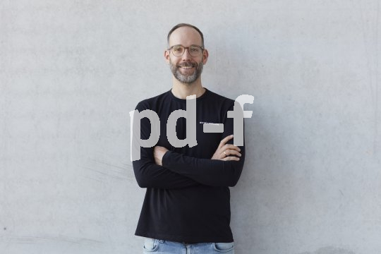 Bild eines Mannes mit Brille, der mit verschränkten Armen vor einer Wand steht