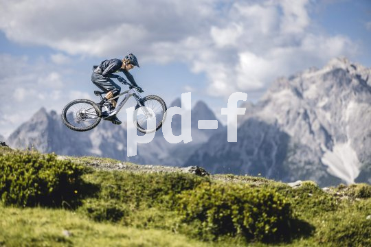 Die Bildunterschrift wird in Bälde eingefügt. Sie können uns aber gern auch per E-Mail oder Telefon kontaktieren, wir helfen gerne weiter. Eine Person springt mit einem E-Mountainbike. Im Hintergrund ist eine alpine Bergkette zu erkennen.