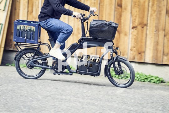Mensch fährt beladenes Cargobike vor Holzwand.