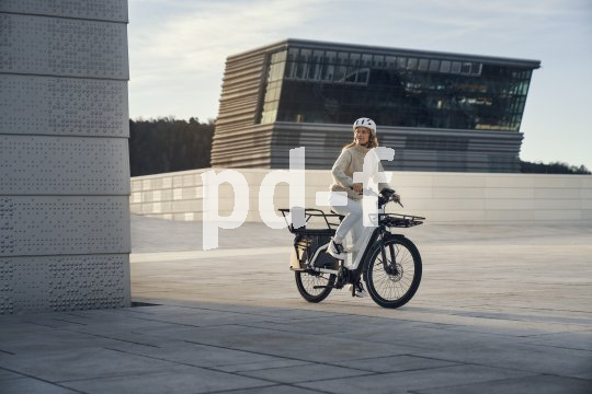 Eine Person auf einem E-Bike mit längerem Gepäckträger vor moderner Architektur.