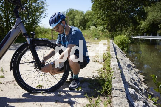 Ein Radfahrer hockt vor einem Fahrrad und löst die Verschraubung des Vorderrades in der Gabel. Unten ist zu sehen, dass der Reifen platt ist.