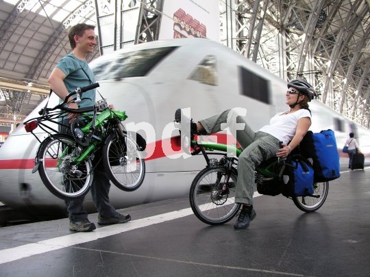 Ob Pendelfahrt oder Radreise: In gefaltetem Zustand passt das Liegerad auch ins Fahrradabteil. Ein Mann mit einem zusammengeklappten Liegerad in der Hand und eine Frau auf einem Liegerad mit Taschen daran auf einem Bahnsteig vor einem weiß-roten Zug.