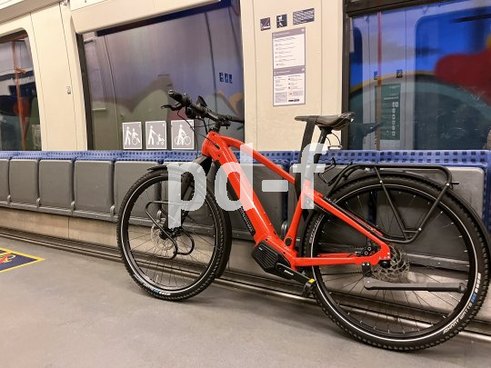 Ein rotes E-Bike lehnt in einem Zug an Klappsitzen.