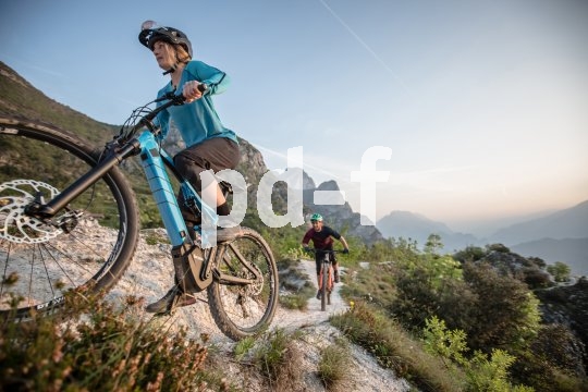 Zwei Personen fahren in einer offenen Gebirgslandschaft auf E-Bikes einen Weg entlang.