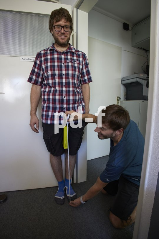 Ein Mann steht in einem Türrahmen und hat ein rechteckiges Objekt zwischen die Beine geklemmt. Ein zweiter Mann misst mit einem Maßband von der Oberkante des Objektes zum Boden.