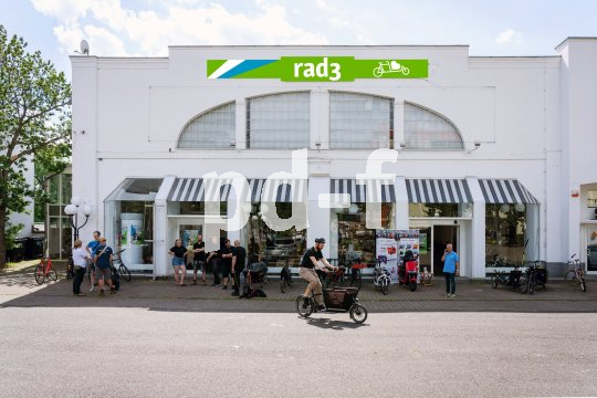 Mehrere Personen stehen vor einem Ladengeschäft mit dem Schriftzug 'rad3'. Ein Mann fährt mit einem Lastenrad los.