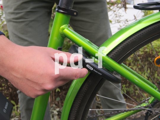 Zum Absichern wird beim Rahmenschloss einfach der Metallring durch die Speichen geschoben. Ein Wegfahren ist somit unmöglich. Eine Person hantiert am Schlüssel eines Rahmenschlosses an einem grünen Fahrrad.