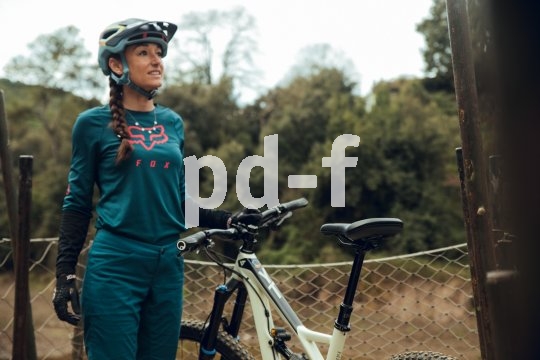 Dieser Sattel besteht aus mehreren Lagen. Eine davon übernimmt die Dämpfung, ähnlich wie bei einer Laufschuhsohle. Eine Frau steht vor einem Zaun vor Bäumen und hält mit der linken Hand ein Mountainbike am Lenker.