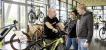 Ein Mann steht mit einem E-Bike in einem Fahrradladen und unterhält sich mit einer Frau und einem Mann.