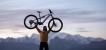 Eine Person steht vor einem Gebirgspanorama und hebt ein E-Bike über den Kopf.