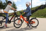 Zwei unterschiedlich große Kinder stehen mit Fahrrädern auf einem Platz und reden miteinander.