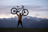 Eine Person steht vor einem Gebirgspanorama und hebt ein E-Bike über den Kopf.