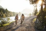 Zwei Personen fahren mit Fahrrädern eine Schotterstraße am Rande eines Flusses.