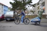 Ein Mann fährt mit einem Fahrrad mit Anhänger durch ein Wohngebiet. Aus dem Anhänger ragt ein verpackter Weihnachtsbaum mit Wimpel an der Spitze heraus