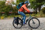 Eine Radfahrerin mit Rucksack und Gepäcktasche am Fahrrad fährt eine Straße mit Kopfsteinpflaster entlang.