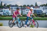 Zwei Kinder fahren Fahrrad. Im Hintergrund ist ein Gewässer und am anderen Ufer Wohnhäuser zu sehen.
