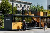 In einer teils offenen Struktur am Straßenrand, die einer Bushaltestelle ähnelt, stellen Menschen Fahrräder ab. Eine Treppe führt auf das Dach, wo Menschen an einem Geländer stehen.