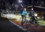 Zwei Personen auf Fahrrädern mit Licht und reflektierenden Elementen an den Fahrradtaschen vor vielen Lichtern einer Großstadt.