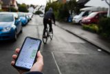 Eine Hand mit einem Smartphone, das einen Stadtplan anzeigt. Unscharf im Hintergrund ist eine Person auf einem Fahrrad in einem Wohngebiet zu sehen.