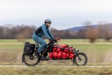 Ein Mann fährt auf einem Lastenrad mit roten Taschen auf der Ladefläche einen Radweg am Feldrand entlang.