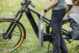 Eine Person steht neben einem E-Bike und hält den länglichen Akku in der Hand, der im Rahmen montiert wird.