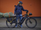 Ein Mann steht neben einem E-Bike mit einem verpackten Weihnachtbaum auf dem Gepäckträger.