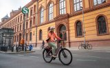 Eine Frau fährt auf einem E-Bike vor einem Gebäude entlang, vor dem ein rundes Schild mit einem weißen S auf grünem Grund steht.