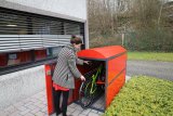 Eine Frau stellt ein Fahrrad in einem roten Metallkasten mit vorne geöffneter Tür ? einer sogenannten Fahrradgarage ? ab.