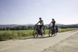 Zwei Personen fahren auf E-Bikes einen betonierten Weg neben einem Feld entlang.