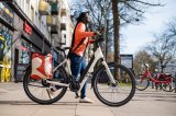 Eine Frau schiebt ein E-Bike über einen Gehweg vor einer Häuserzeile mit Geschäften.