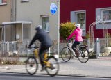 Zwei Personen fahren in dieselbe Richtung auf Fahrrädern durch die Stadt ? eine auf der Fahrbahn und eine auf dem geteilten Fuß- und Radweg.