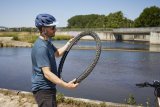 Ein Radfahrer steht vor einem Gewässer und begutachtet einen Fahrradreifen, den er mit beiden Händen vor sich hält.