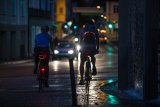 Zwei Personen fahren nachts auf Fahrrädern durch eine Innenstadt. An ihren Fahrrädern sind Rücklichter zu sehen und an ihren Rucksäcken und Taschen leuchtende oder reflektierende Elemente.