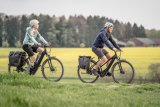 Eine Frau und ein Mann fahren auf E-Bikes zwischen Feldern und Wiesen entlang.