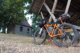 Ein mit Gepäck beladenes, orangefarbenes Mountainbike lehnt an einem hölzernen Unterstand auf einer Wiese vor einer Kapelle.