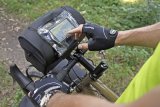 Eine Person tippt mit dem Finger auf das Display eines Smartphones oder Navigationsgeräts in einer Klarsichthülle auf dem Deckel einer Lenkertasche eines Fahrrades.