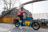 Eine Frau fährt auf einem blauen Lastenrad mit einem gelben Getränkekasten auf der Ladefläche über eine Brücke in der Stadt.