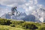 Eine Person springt mit einem E-Mountainbike. Im Hintergrund ist eine alpine Bergkette zu erkennen.