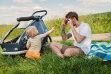 Ein Mann sitzt auf einer Wiese neben einem Fahrradanhänger und fotografiert mit einer bunten Kamera ein kleines Kind.