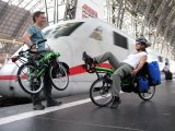 Ein Mann mit einem zusammengeklappten Liegerad in der Hand und eine Frau auf einem Liegerad mit Taschen daran auf einem Bahnsteig vor einem weiß-roten Zug.