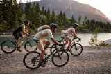 Drei Personen fahren auf Gravelbikes auf einem Schotterweg am Ufer eines Gewässers in den Bergen entlang.